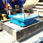 Construction workers carefully installing a blue Hercules POT Bearing into position within a concrete formwork on a construction site.