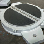 A heavy-duty, silver-colored structural bearing with thick plates and large bolts, viewed in a manufacturing facility.