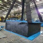 An industrial crane carefully lifting a large elastomeric bearing pad in a factory.